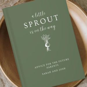 May include: A green hardcover book with the text "a little SPROUT is on the way" and "ADVICE FOR THE FUTURE PARENTS SARAH AND JOSH" in white. The book is on a wooden plate.
