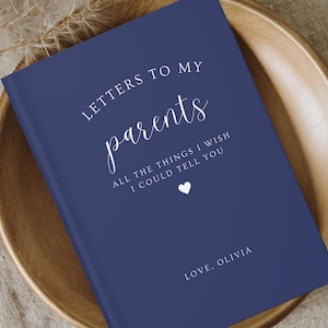 Puede incluir: Un diario azul marino con el texto "LETTERS TO MY parents ALL THE THINGS I WISH I COULD TELL YOU" y un pequeño corazón blanco. El texto "LOVE, OLIVIA" está en la parte inferior del diario.