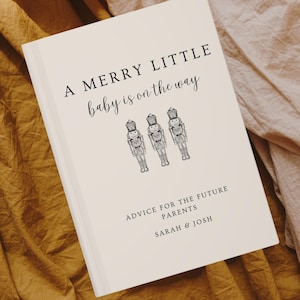 Peut inclure: Un livre blanc avec le texte "A Merry Little baby is on the way" et trois illustrations de casse-noisettes. Le texte "Advice for the Future Parents Sarah & Josh" est en bas du livre.
