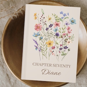 May include: A cream-coloured journal with a floral design featuring pink, yellow, blue, and purple flowers. The text "CHAPTER SEVENTY Diane" is printed on the front cover.