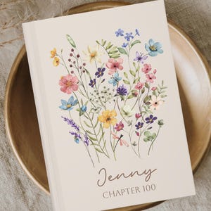 May include: A hardcover journal with a floral design featuring pink, blue, yellow, and purple flowers. The cover is cream-colored with the name "Jenny" and "Chapter 100" printed in a script font.