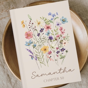 May include: A hardcover book with a floral design featuring pink, blue, yellow, and purple flowers. The book cover is cream-colored with the name "Samantha" and "CHAPTER 50" printed on it. The book is resting on a wooden plate.