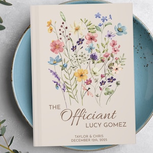May include: A white book with a floral design on the cover. The text "The Officiant Lucy Gomez" is printed on the cover, along with the names "Taylor & Chris" and the date "December 12th, 2025".