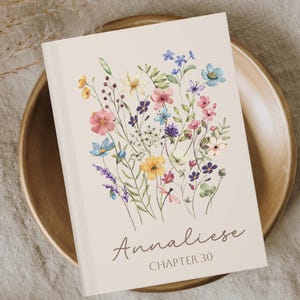 May include: A book cover with a floral design featuring pink, blue, yellow, and purple flowers. The title "Annaliese" is written in a cursive font, with "CHAPTER 30" below it. The book is resting on a wooden plate.