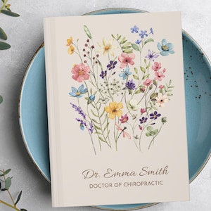 May include: A beige journal with a watercolor floral design featuring pink, blue, yellow, and purple flowers. The journal is inscribed with "Dr. Emma Smith, DOCTOR OF CHIROPRACTIC" in elegant script.