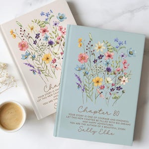 Puede incluir: Dos libros de tapa dura con ilustraciones florales en las portadas. El libro azul presenta el texto "Chapter 80, Your story is one of courage and kindness, let this new chapter be filled with big dreams and happy adventures, you are the author of a beautiful story, Sally Elke."