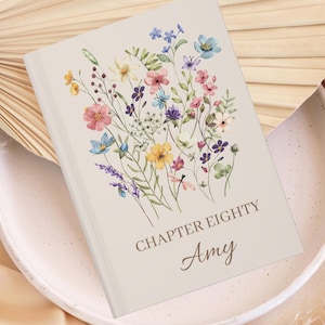Puede incluir: Un diario beige con un diseño floral que presenta una variedad de flores coloridas. El texto "CHAPTER EIGHTY Amy" está impreso en la portada.