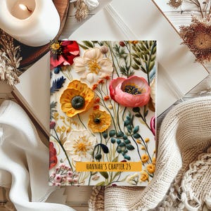 May include: A journal with a floral design featuring red, yellow, and pink flowers. The journal cover has the text "HANNAH'S CHAPTER 25" on a yellow banner. The journal is open on a white surface, with a lit candle and a knitted blanket in the background.