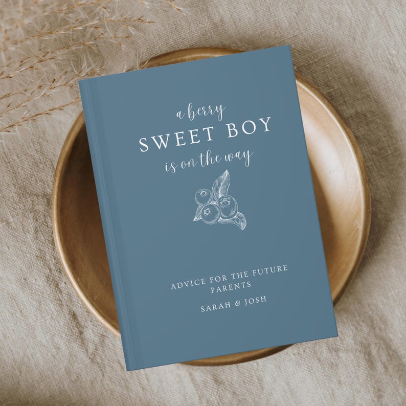 K&ouml;nnte beinhalten: Ein blaues Notizbuch mit dem Text "a berry SWEET BOY is on the way" und "ADVICE FOR THE FUTURE PARENTS SARAH & JOSH". Das Cover zeigt eine Blaubeer-Illustration. Das Notizbuch liegt in einer Holzschale.