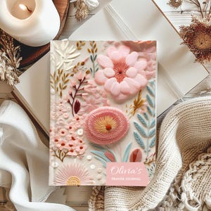 May include: A prayer journal with a floral design. The cover features a variety of pink flowers and leaves in different textures and shades. The journal has the text "Olivia's Prayer Journal" on a pink label.