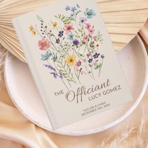 May include: A beige book with a floral design on the front cover. The text "THE Officiant" is written in brown cursive lettering. Below the text is the name "LUCY GOMEZ" in brown lettering. Below the name is the text "TAYLOR & CHRIS DECEMBER 12th, 2025" in brown lettering.