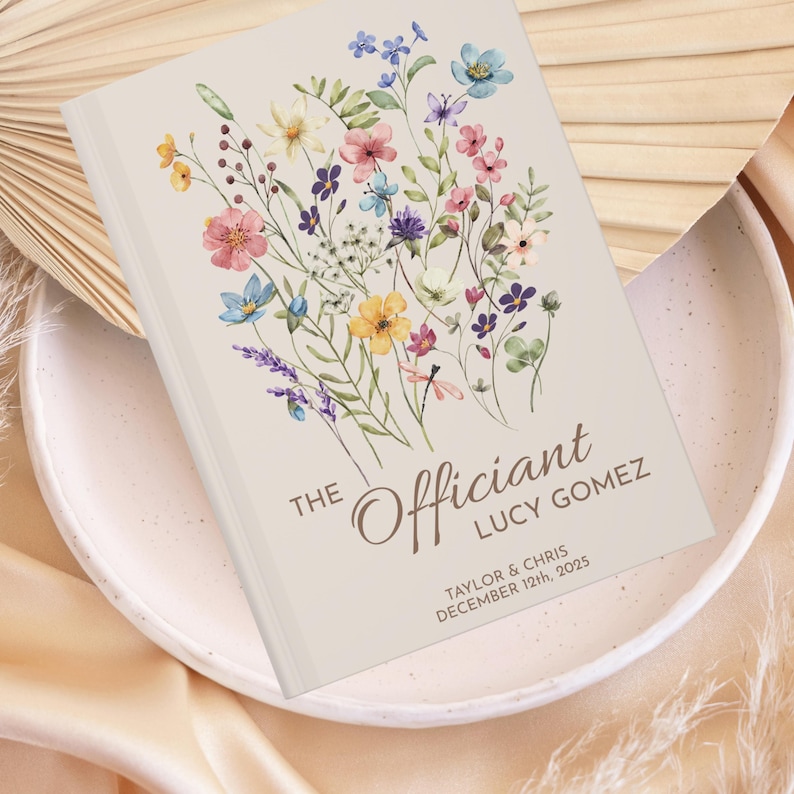 May include: A beige book with a floral design on the front cover. The text "THE Officiant" is written in brown cursive lettering. Below the text is the name "LUCY GOMEZ" in brown lettering. Below the name is the text "TAYLOR & CHRIS DECEMBER 12th, 2025" in brown lettering.