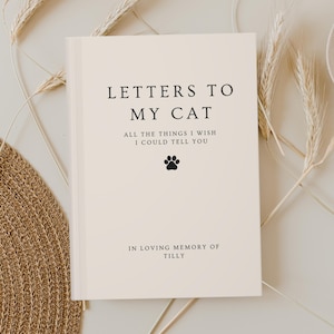 Op de afbeelding: Een crèmekleurig boek met de titel "LETTERS TO MY CAT" met zwarte tekst. Het boek bevat de tekst "ALL THE THINGS I WISH I COULD TELL YOU" en "IN LOVING MEMORY OF TILLY". Een zwarte pootafdruk is ook zichtbaar.