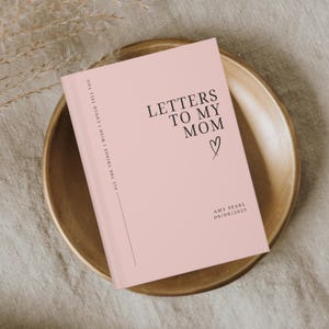 May include: A pink journal titled "Letters to My Mom" with a heart symbol. The journal includes the text "All the things I wish I could tell you" and the name "Amy Pearl" with a date. The journal is on a wooden plate.