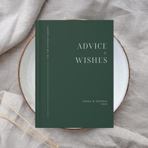May include: A dark green book with the text "Advice + Wishes" in white on the front cover. The book is on a white plate and has the names "Jenna & Thomas" and the year "2024" printed below the title. The book is titled "For the Future Parents".