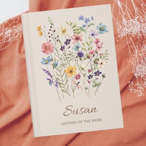 May include: A white journal featuring a watercolour floral design in shades of pink, yellow, blue, and green. The text "Susan Mother of the Bride" is printed in brown at the bottom.