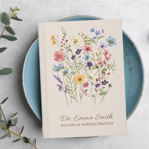 Può includere: Un diario beige con un design floreale ad acquerello con fiori rosa, blu, gialli e viola. Il diario è personalizzato con il testo "Dr. Emma Smith, DOCTOR OF NURSING PRACTICE" in un carattere corsivo.
