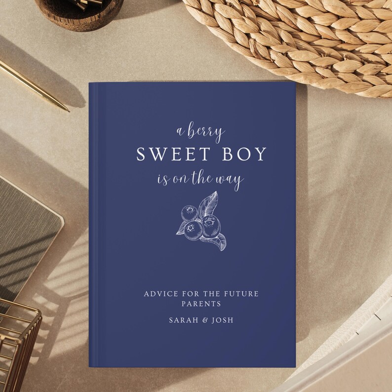 K&ouml;nnte beinhalten: Ein marineblaues Hardcover-Buch mit dem Text "a berry SWEET BOY is on the way" und "ADVICE FOR THE FUTURE PARENTS SARAH & JOSH". Eine wei&szlig;e Illustration von Blaubeeren ist ebenfalls abgebildet.