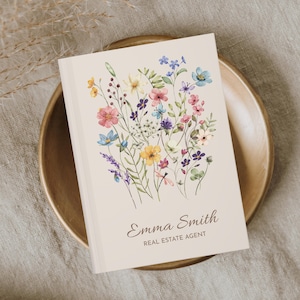 Puede incluir: Un cuaderno crema con un diseño floral en acuarela. La portada presenta un ramo de flores rosas, azules, amarillas y moradas. El nombre "Emma Smith" y el título "REAL ESTATE AGENT" están impresos.