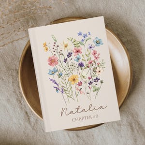 May include: A cream-coloured journal with a watercolour floral design featuring pink, blue, yellow, and purple flowers. The name "Natalia" and "CHAPTER 40" are written in brown script. The journal is on a wooden plate.