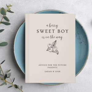 K&ouml;nnte beinhalten: Ein cremefarbenes Buch mit dem Text "a berry SWEET BOY is on the way" und einer Blaubeer-Illustration. Darunter steht "ADVICE FOR THE FUTURE PARENTS" und die Namen "SARAH & JOSH". Das Buch liegt auf einem blauen Teller.