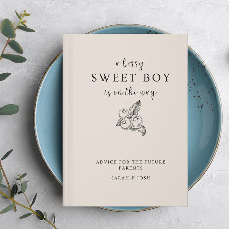 K&ouml;nnte beinhalten: Ein cremefarbenes Buch mit dem Text "a berry SWEET BOY is on the way" und einer Blaubeer-Illustration. Darunter steht "ADVICE FOR THE FUTURE PARENTS" und die Namen "SARAH & JOSH". Das Buch liegt auf einem blauen Teller.