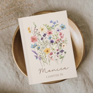 May include: A cream-colored book cover featuring a watercolor floral design in pink, blue, yellow, and purple. The name "Monica" is written in cursive, with "CHAPTER 70" below. The book rests on a wooden plate.