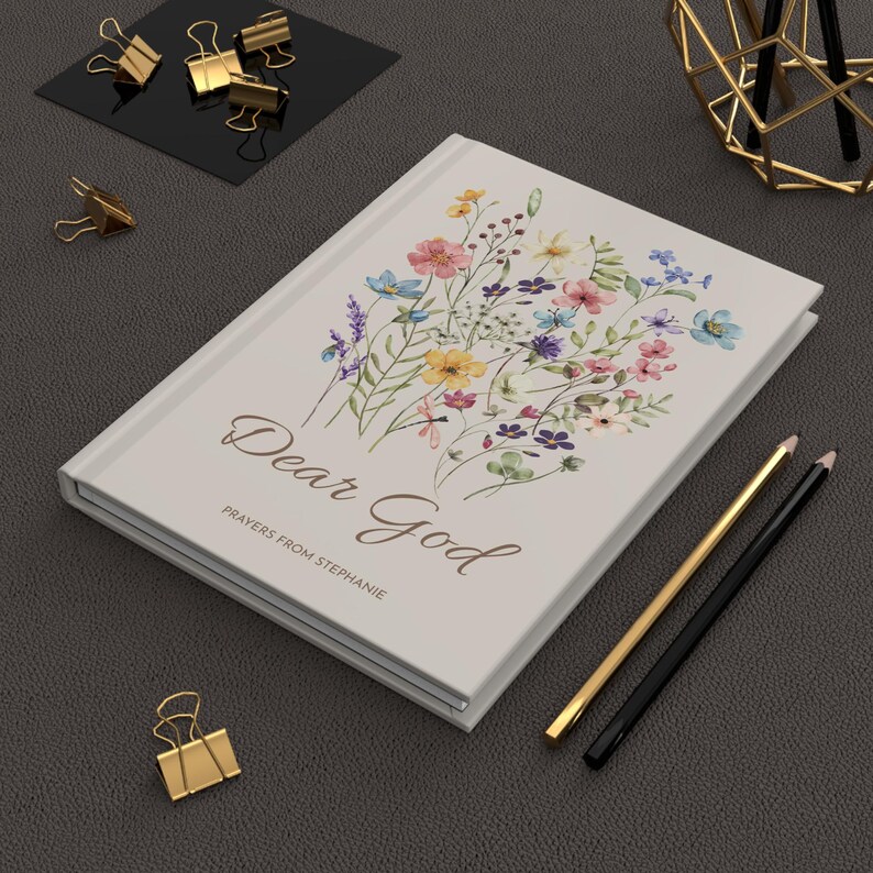 May include: A cream-coloured journal with the words "Dear God" and floral illustrations. The journal also has the text "Prayers from Stephanie". Gold and black pencils and gold binder clips are also visible.