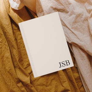 Puede incluir: Un cuaderno blanco con las iniciales "JSB" impresas en negro en la portada. El cuaderno est&aacute; sobre un fondo de tela marr&oacute;n y rosa.