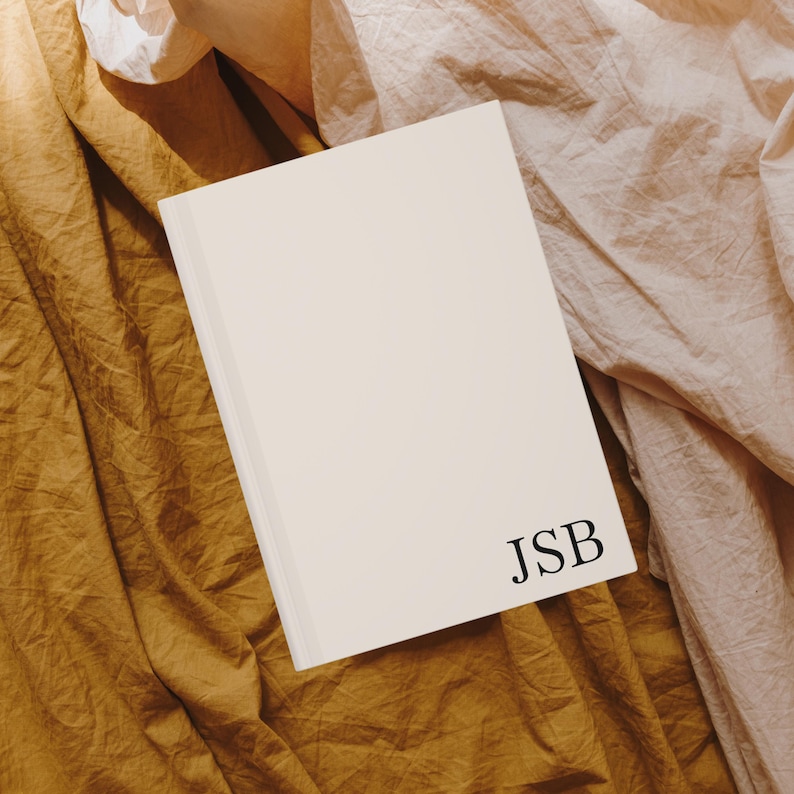 Puede incluir: Un cuaderno blanco con las iniciales "JSB" impresas en negro en la portada. El cuaderno est&aacute; sobre un fondo de tela marr&oacute;n y rosa.