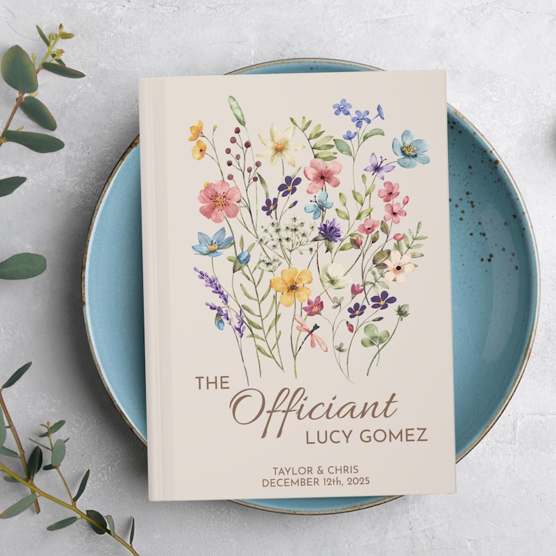 May include: A white book with a floral design on the cover. The text "The Officiant Lucy Gomez" is printed on the cover, along with the names "Taylor & Chris" and the date "December 12th, 2025".