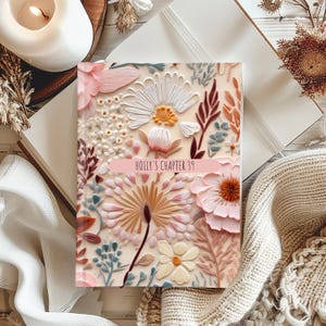 Puede incluir: Un diario floral con un fondo crema y una variedad de flores en relieve en rosa, blanco y beige. La portada del diario tiene el texto "HOLLY'S CHAPTER 39" en una pancarta rosa. El diario está abierto sobre una superficie blanca.