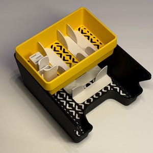 May include: A two-tiered drawer organizer featuring a black base and a yellow top tray. Both levels contain white dividers and a black and white chevron patterned liner. The structure is rectangular with a cutout section on the front side.