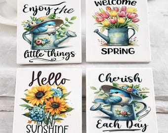 May include: A set of four 4x4 inch ceramic coasters with floral and bird designs. Each coaster features a different inspirational message: "Enjoy the little things," "Welcome Spring," "Hello Sunshine," and "Cherish Each Day."