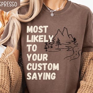 May include: Espresso-colored t-shirt with the text "MOST LIKELY TO YOUR CUSTOM SAYING" in white block letters. A black line drawing of mountains, trees, and a road is also featured on the shirt.