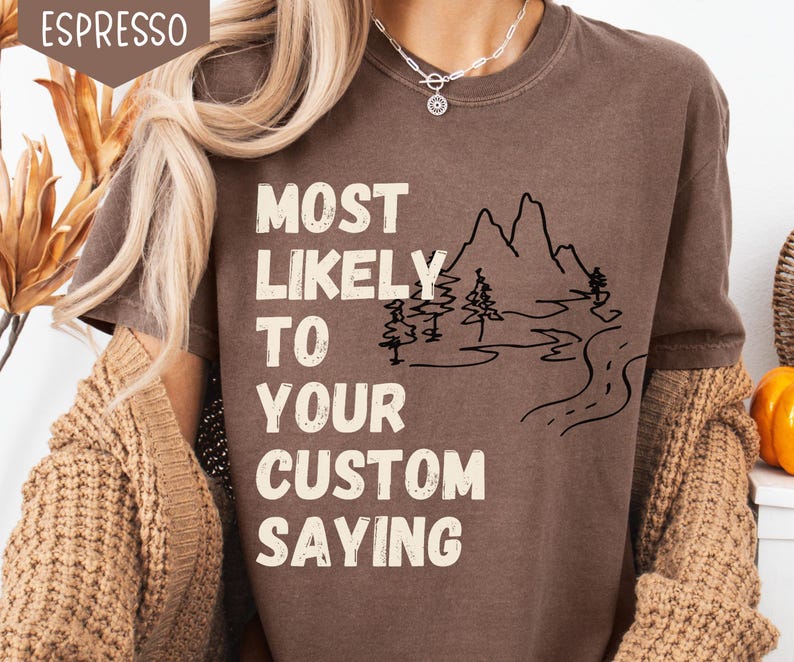 May include: Espresso-colored t-shirt with the text "MOST LIKELY TO YOUR CUSTOM SAYING" in white block letters. A black line drawing of mountains, trees, and a road is also featured on the shirt.