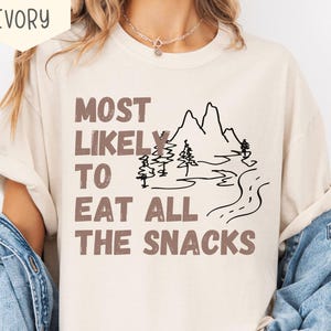 May include: Ivory-colored t-shirt with the text "MOST LIKELY TO EAT ALL THE SNACKS" in brown letters. A black line drawing of mountains, trees, and a road is also featured on the shirt. The shirt has short sleeves.