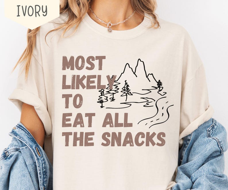 May include: Ivory-colored t-shirt with the text "MOST LIKELY TO EAT ALL THE SNACKS" in brown letters. A black line drawing of mountains, trees, and a road is also featured on the shirt. The shirt has short sleeves.