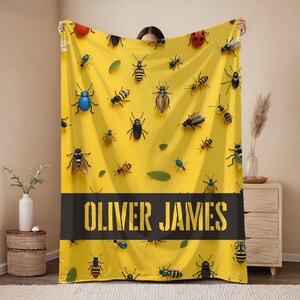 May include: A yellow blanket featuring a pattern of various insects and green leaves. A black stripe across the bottom displays the name "OLIVER JAMES" in a bold, stencil-style font. The blanket is held up by a person.