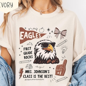 May include: Ivory-colored t-shirt with a graphic of an eagle wearing sunglasses, the word "EAGLES", and the text "FIRST GRADE ROCKS". The shirt also includes the text "MRS. JOHNSON'S CLASS IS THE BEST!" and various doodles.