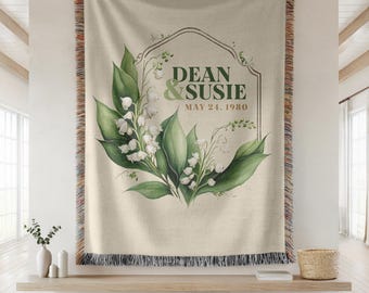 May include: A cream-colored woven tapestry with a fringe border. The tapestry features a floral design with green leaves and white flowers. The text "DEAN & SUSIE MAY 24, 1980" is printed in a brown frame.