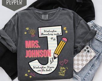 May include: A dark gray long-sleeve shirt with a colorful graphic design. The design includes the text "MRS. JOHNSON" and "Washington Elementary rocks!" along with a pencil, stars, and hearts. The shirt also has the text "Washington Tigers are the best!" and "You've Got This!"