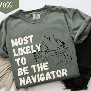 May include: A moss green t-shirt with the text "MOST LIKELY TO BE THE NAVIGATOR" in white block letters. A black line drawing of mountains, trees, and a road is next to the text. The shirt is paired with black denim jeans.