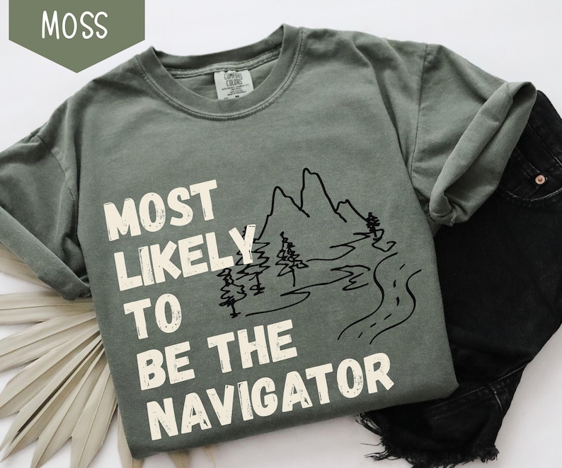 May include: A moss green t-shirt with the text "MOST LIKELY TO BE THE NAVIGATOR" in white block letters. A black line drawing of mountains, trees, and a road is next to the text. The shirt is paired with black denim jeans.