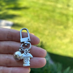 May include: A silver-toned metal keychain featuring a lobster clasp and two charms. One charm is a spherical soccer ball, and the other is a jersey-shaped charm with the word SOCCER and a small soccer ball graphic embossed on the front surface.
