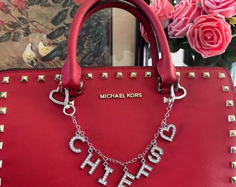May include: A red Michael Kors handbag featuring gold-tone studs along the edges and a silver-tone chain charm attached to the handles. The charm displays the word CHIEFS in rhinestone-encrusted letters, accented with small silver heart charms.