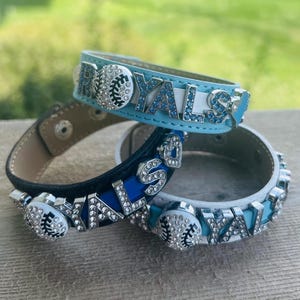 May include: Three leather bracelets with rhinestones and baseball charms. The bracelets are blue, black, and white. The blue bracelet says "ROYALS" in rhinestones. The black bracelet has a baseball charm and rhinestones. The white bracelet has a baseball charm and rhinestones and says "ROYALS" in rhinestones.