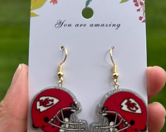 May include: A pair of dangle earrings featuring red football helmet charms with white and black details and a silver glitter finish. The earrings hang from gold-toned hooks on a white display card with floral accents and the text You are amazing.