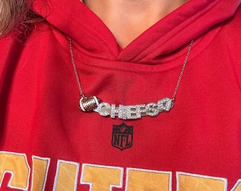 May include: A silver necklace featuring a football charm and the word "CHIEFS" spelled out in rhinestones. The necklace is worn by a person wearing a red hoodie with the NFL logo and the word "CHIEFS" embroidered on the front.