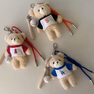 May include: Three plush teddy bear keychains, each wearing a different baseball team jersey. The bears are holding miniature baseball bats and baseballs. The jerseys are for the San Francisco Giants, Los Angeles Angels, and Los Angeles Dodgers.